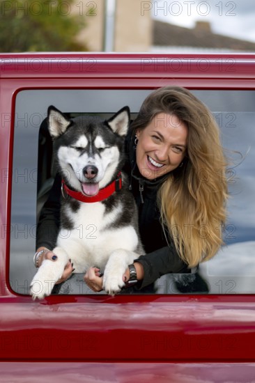 A joyful woman with long hair leans out of a red car window, holding a happy husky dog wearing a red collar, capturing a moment of pure happiness and companionship