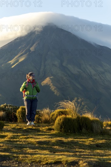 A woman wearing hiking gear stands thoughtfully in front of Mount Taranaki, New Zealand, as the morning sunlight casts a warm glow over the landscape