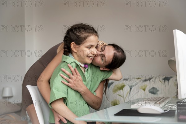 Young disabled girl with a tracheostomy, being embraced and kissed by sister while using desktop computer at home