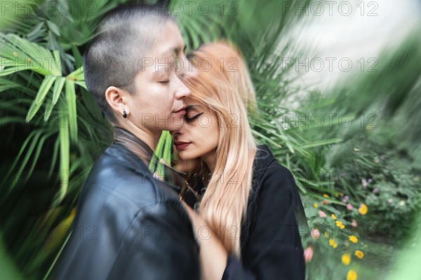 A lesbian couple shares a tender, intimate moment surrounded by vibrant greenery, symbolizing love and connection. The lush foliage adds a serene backdrop to their embrace
