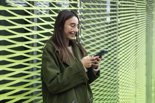 A woman wearing a stylish green coat is smiling as she uses her smartphone She stands near a modern, artistic green wall, which complements her outfit perfectly