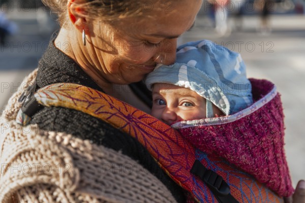 A tender moment between a mother and her baby in a colorful carrier. The warm sunlight illuminates their joyful smiles, capturing the essence of love and connection