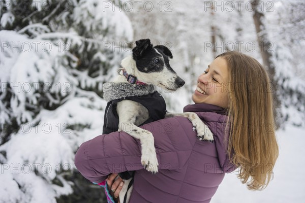 A woman in a purple jacket holds her dog lovingly in a snowy forest. Her smile and the snowy scene capture the joy and warmth of a winter day spent outdoors