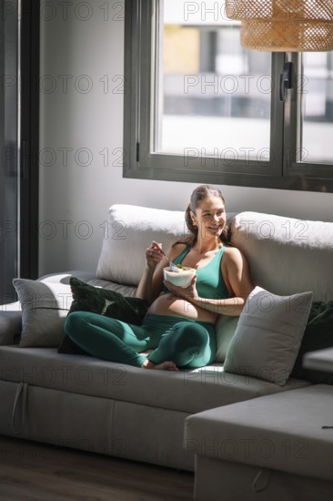 Pregnant woman in a green yoga outfit relaxes on a sofa, enjoying a healthy bowl of fruit. Sunlight pours in, highlighting her peaceful expression and the cozy atmosphere