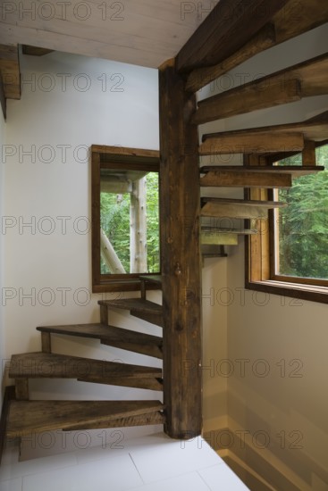 Brown stained cherry wood spiraling staircase with thin flat wooden steps attached to pine wood post inside Scandinavian hybrid style log cabin home, Quebec, Canada
