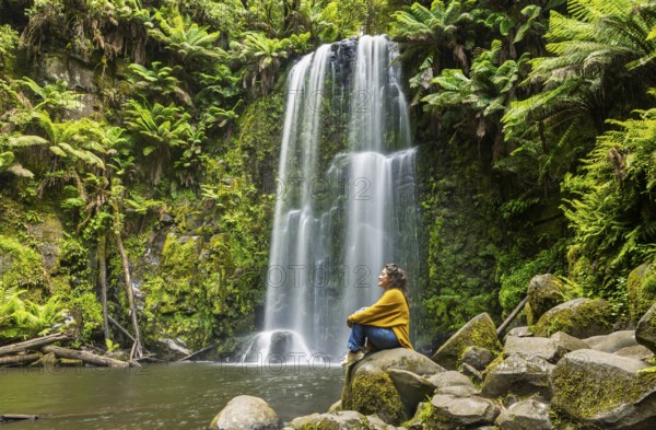 A woman sits peacefully on moss covered rocks near a waterfall surrounded by lush green ferns in the Great Ocean Road, Australia. Serenity and nature combine in this tranquil forest setting
