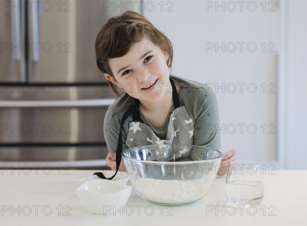 A young child in an apron is smiling while preparing to bake in a modern kitchen. The scene features a glass bowl of flour and a glass of water nearby