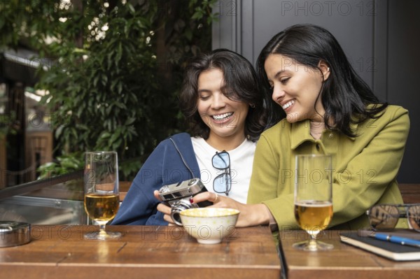 A joyful lesbian couple enjoys a sunny afternoon together, sharing moments with a vintage camera. They are seated in a cozy outdoor setting with drinks and smiles