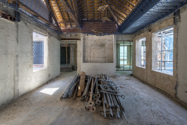 A spacious room in an unfinished building shows construction materials on the floor, open windows, and a safety net on the ceiling. Light filters through, highlighting the ongoing renovation work