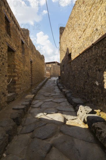 Long narrow street made of large grey smooth and worn lava stones with sidewalk plus remnants of old terracotta brick and stone wall structures and residential buildings at the ancient ruins of Pompeii, Campania region, Italy