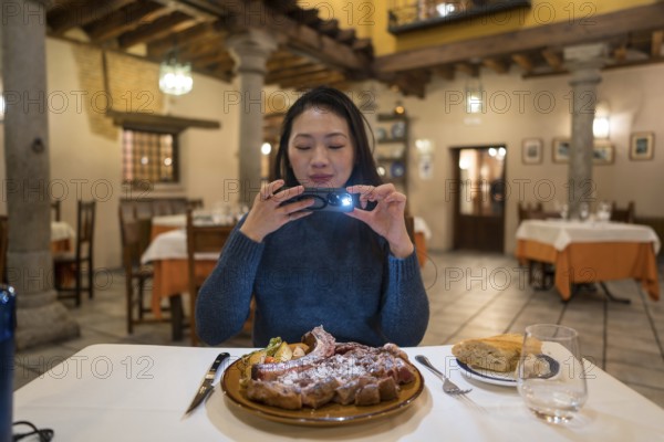An Asian woman captures a photo of an Avila T-bone steak before dining in a restaurant with a warm and rustic interior, showcasing a delightful culinary experience