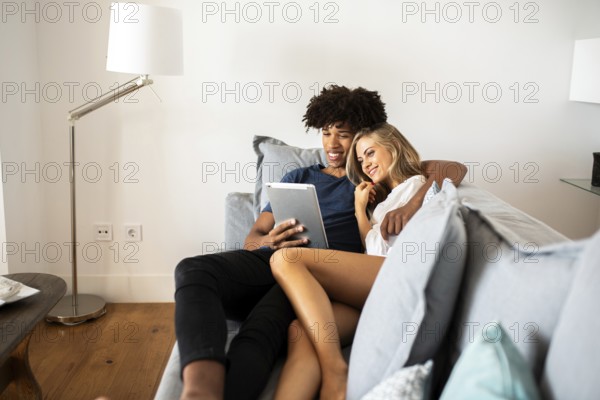 A young couple relaxes on a cozy couch, sharing a tablet. The scene captures a moment of joy and connection, with a modern lamp and bright, inviting atmosphere