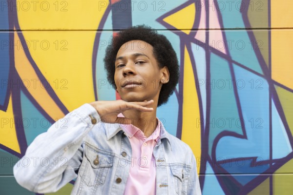 A confident non-binary person stands proudly in front of a vibrant graffiti mural. They wear a light denim jacket over a pink shirt, showcasing style and individuality against the colorful background