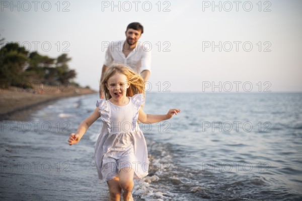 A young girl runs barefoot through the shallow waves of an Italian beach, smiling with joy as her father follows behind. They enjoy a playful and carefree summer evening together