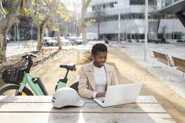 A professional black woman sits at a table in an urban park, working on her laptop Nearby, a bicycle and helmet suggest a modern, eco-friendly lifestyle