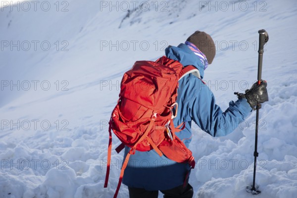 A skier with a red backpack explores a winter landscape during a ski touring adventure. The snow-covered mountains provide a serene backdrop, highlighting the thrill of outdoor exploration