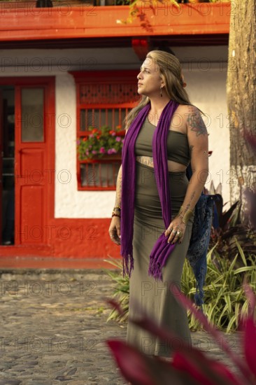 An elegant woman with tattoos, purple scarf and tight green dress walks down a vibrant and colorful street. Her casual yet elegant look complements the urban setting, reflecting individuality and exploration.