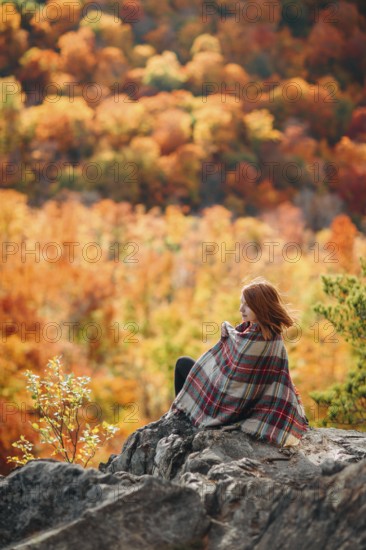A woman sits on a rocky outcrop, wrapped in a plaid blanket, savoring the vibrant hues of autumn in a Quebec forest. The scene captures the essence of tranquility and natural beauty