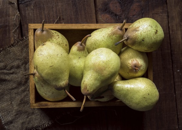 A top view of ripe pears stacked in a wooden box, placed on a dark wooden surface. The rustic setting and vibrant green hues highlight freshness and simplicity