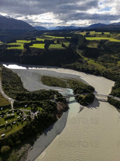 Aerial view captures the diverse landscape of Canterbury, New Zealand, highlighting winding rivers, lush farmlands, a bridge, and distant mountains under a cloudy sky