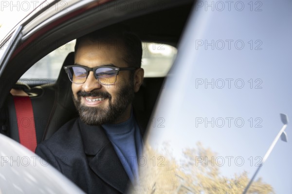 Cheerful Indian businessman looks away while smiling in his car, dressed in winter attire, reflecting a joyful commuting experience
