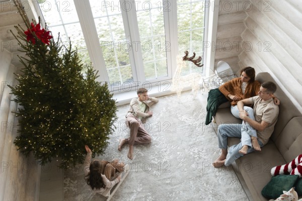 A family celebrates Christmas in a cozy room adorned with a large, lit Christmas tree. A couple sits on a couch two children play nearby, a girl reaches up towards the tree, and a boy lies on the floor looking away