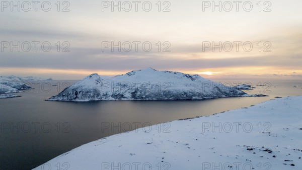 A breathtaking aerial view captures a Nordic winter landscape with a tranquil snowy island surrounded by calm waters under a soft, pastel sunset sky, evoking serene beauty, Kvaløya, Troms, Norway