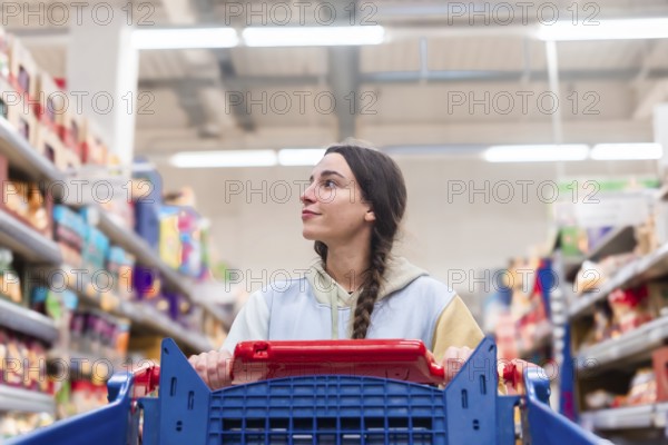 A woman with a braided ponytail glances upwards while pushing a red shopping cart in a supermarket aisle