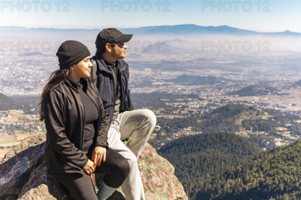 Two friends are seated on a mountain rock, gazing at a stunning panoramic view They are bundled up against the crisp mountain air on a clear day, admiring the scenery