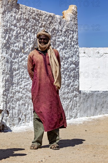 A man in traditional Tuareg clothing stands in front of a sunlit, plastered wall, Local people in the Sahara in Algeria