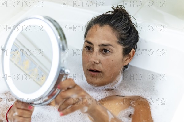 A woman lies in a bubble bath, holding a round mirror. The scene captures a serene moment of self reflection at home, with a focus on relaxation and personal care