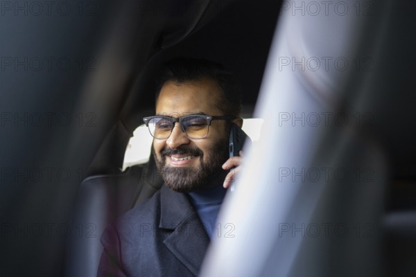 A smiling Indian man dressed in a winter coat and sweater, talking on a mobile phone while sitting in a car, looking away from the camera