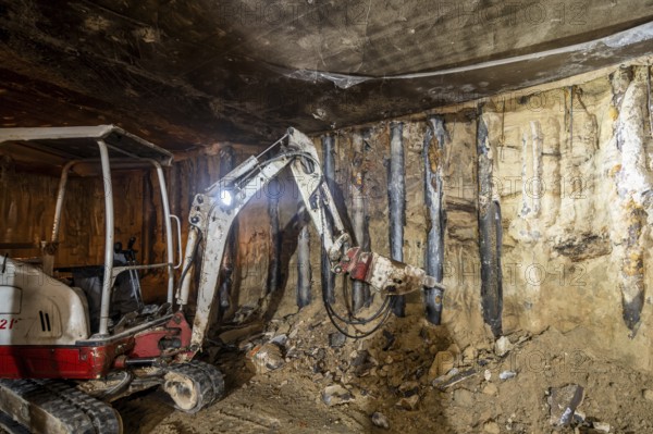 An excavator operates in an underground construction site, tearing through rock and earth. The dim lighting highlights the machine's powerful build and the raw texture of the walls