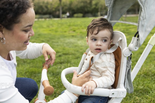 A loving mother bends down to interact with her baby sitting in a stroller. They are enjoying a sunny day at the park, surrounded by lush green grass and peaceful scenery