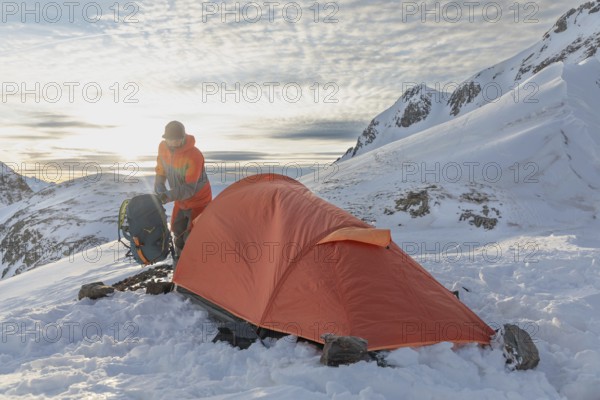 A ski tourer sets up near a bright orange tent amidst snowy mountains at sunrise, capturing the spirit of adventure and outdoor exploration in a stunning winter landscape