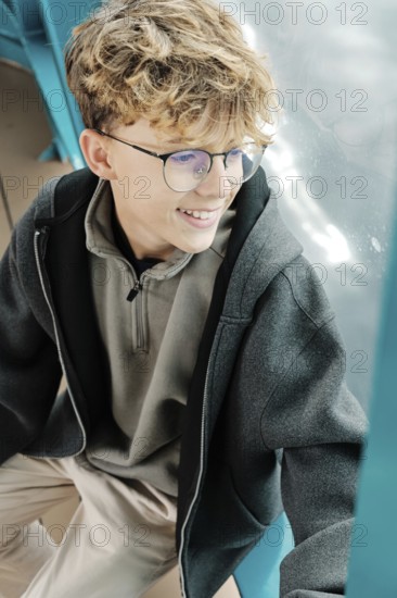 A curly-haired teenager with glasses sits by a window, smiling as he enjoys a ski resort in the Pyrenees. His relaxed posture and comfortable clothing create a moment of serene reflection