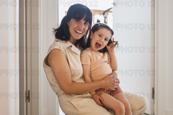 A joyful young mother lovingly embraces her playful toddler daughter in a bright home environment. They share a moment of happiness and warmth, reflecting family love