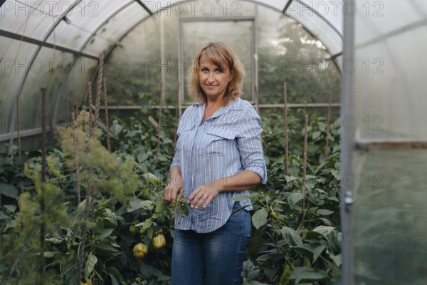 A woman in casual attire stands amidst lush plants inside a greenhouse. She tends to the greenery, embodying dedication to sustainable agriculture and horticulture