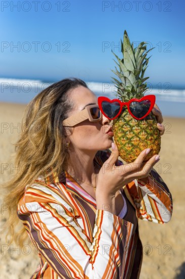 Woman playing with a pineapple wearing red heart sunglasses, standing on a sandy beach by the ocean, embodying summer fun and exotic vacation vibes under a clear blue sky