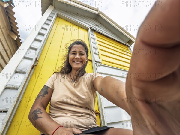 A joyful woman takes a close-up selfie in front of a vibrant yellow Brighton bathing box in Melbourne, Australia. She is smiling warmly, enjoying a sunny beach day