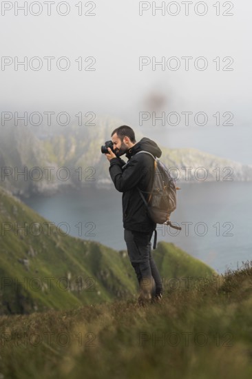A photographer with a backpack captures the beauty of Norway's coastal cliffs, surrounded by lush greenery and a misty atmosphere Perfectly embodies outdoor adventure and creativity