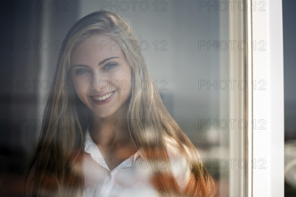 A young woman smiles gently through a glass window, captured in soft natural light. Her relaxed expression and casual attire create a serene and inviting atmosphere