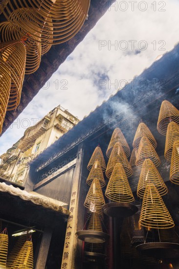 Spiral incense cones hang within the dimly lit interior of a Taoist temple in Macau, emitting fragrant smoke, against a backdrop of traditional architecture
