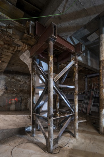 A metal support structure stands firm in a dimly lit construction site. Exposed beams, wooden planks, and dust create an industrial atmosphere. Overhead cables add to the raw setting
