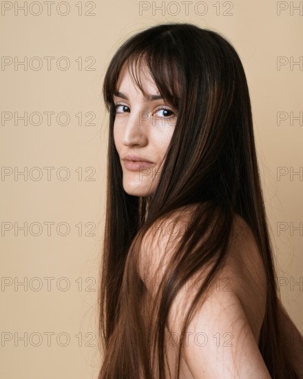 A portrait of a young woman with long brown hair, looking at camera over her shoulder on a beige background in a studio. Her gaze is subtle, adding depth to the image