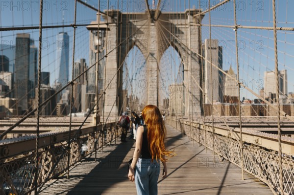 A woman with long hair walks on the iconic Brooklyn Bridge in New York City, capturing the stunning skyline under a clear. The historic structure and vibrant city life create a picturesque scene