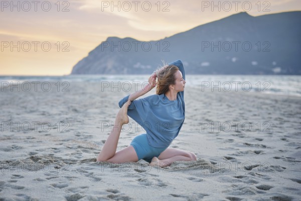 A woman does Eka Pada Rajakapotasana on a sandy beach while the sun sets over the water and hills in the background