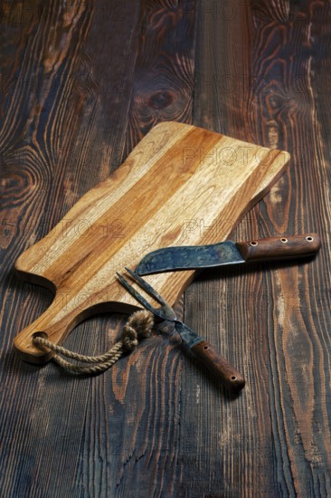 Shabby wooden table with empty cutting board and old forged knife and fork for barbecue
