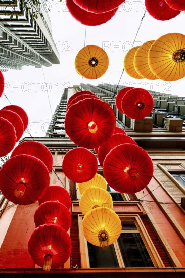 From below radiant red and yellow lanterns hang between skyscrapers in Hong Kong, celebrating Chinese New Year. The festive decorations create a vibrant canopy against the urban backdrop, symbolizing prosperity and good fortune