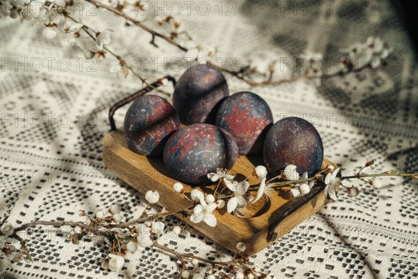 Cosmic-painted Easter eggs displayed on a rustic wooden tray, adorned with delicate white spring blossoms, set against a patterned crochet tablecloth background